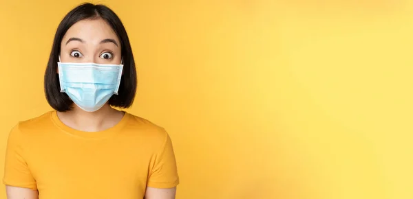 Covid-19 en medisch concept. Close-up portret van aziatische vrouw in het gezicht masker, op zoek verrast en verbaasd over nieuws, staande over gele achtergrond — Stockfoto