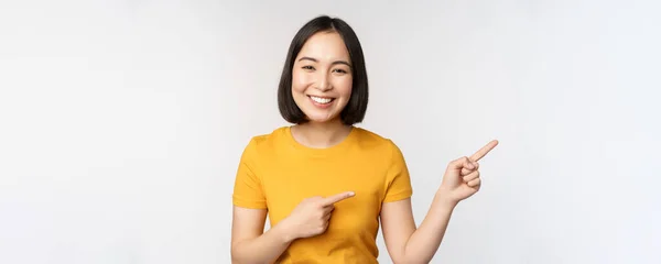 令人惊讶的年轻亚洲女人，把广告放在一边，用手指指向促销文字、品牌标识，在白色背景下快乐地站着 — 图库照片