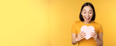 Romantizm ve sevgililer günü. Büyük bir kalp kartı tutan, gülümseyen, sarı arka planda duran mutlu, güzel Asyalı kadın.