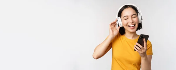 Linda chica japonesa en los auriculares, mirando el teléfono móvil y sonriendo, utilizando la aplicación de música en el teléfono inteligente, de pie sobre fondo blanco — Foto de Stock