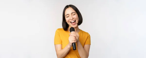 Feliz chica asiática cantando y divirtiéndose, sosteniendo el micrófono en el karaoke, de pie en camiseta amarilla sobre fondo blanco —  Fotos de Stock