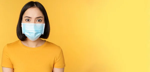 Портрет шокованої жінки, яка виглядає стурбованою і вражений камерою, одягнена в медичну маску коварід-19, стоячи проти жовтого фону. — стокове фото