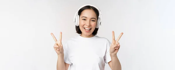 Happy asian woman wearing headphones and smiling, showing peace v-sign, listening music, standing in tshirt over white background — Stock Photo, Image