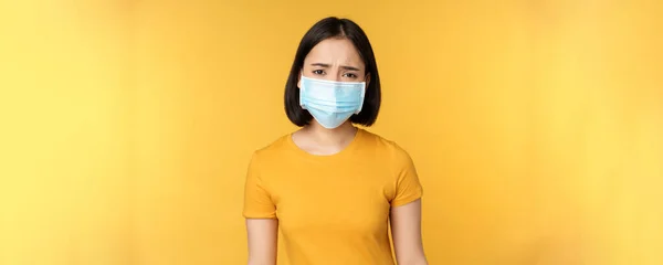 Εικόνα λυπημένης και απογοητευμένης Ασιάτισσας με ιατρική μάσκα, συνοφρύωμα και έκφραση δυσαρέσκειας, να στέκεται απέναντι σε κίτρινο φόντο — Φωτογραφία Αρχείου