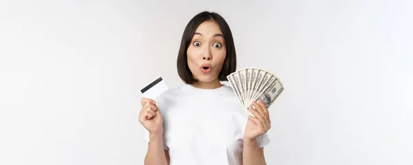 Ritratto di donna asiatica in possesso di denaro, dollari e carta di credito, dall'aspetto impressionato e stupito, in piedi in maglietta su sfondo bianco — Foto Stock