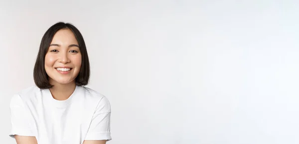 Hermosa chica coreana sonriendo, dientes blancos, con un aspecto encantador en la cámara, de pie en camiseta blanca sobre fondo del estudio —  Fotos de Stock