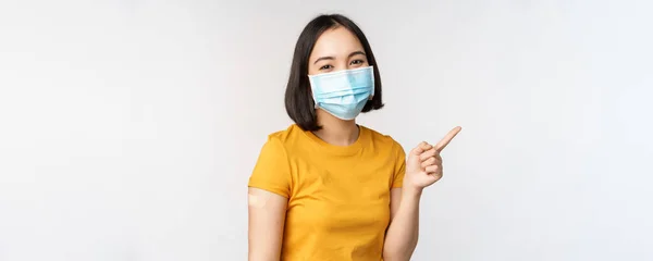 Covid-19, vaccinations- och sjukvårdskoncept. Porträtt av söt asiatisk flicka i medicinsk mask, har band stöd på axeln efter coronavirus vaccin, står över vit bakgrund — Stockfoto