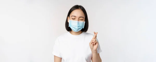Ковид-19, медицина и медицинская концепция. Изображение азиатки в маске медицинского лица, скрещенные пальцы, молящиеся, загадывающие желание и улыбающиеся, стоящие на белом фоне — стоковое фото