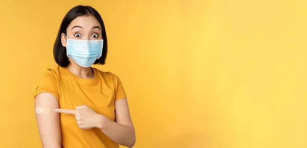Vaccination from covid and health concept. Excited asian woman in medical face mask, pointing finger at shoulder with band aid, standing over yellow background — Stock Photo, Image