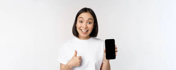 Mujer joven entusiasta mostrando el pulgar hacia arriba y la pantalla del teléfono móvil, de pie en camiseta sobre fondo blanco —  Fotos de Stock