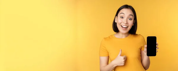 Excited asian woman showing mobile phone screen, thumbs up, like smth good, recommending smartphone application, standing over yellow background — Stock Photo, Image