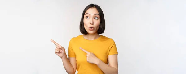 Förvånad asiatisk kvinna pekar och tittar vänster på kopia utrymme, PR-försäljning, visar reklam med imponerade ansiktsuttryck, står över vit bakgrund — Stockfoto