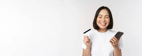 Handla på nätet. Glad asiatisk kvinna med kreditkort och smartphone app, betala på webbplatsen via mobiltelefon, vit bakgrund — Stockfoto