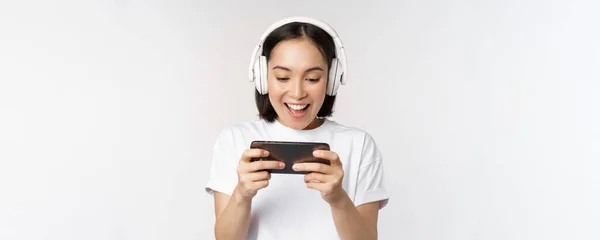 Glückliche Asiatin mit Kopfhörer, Blick aufs Smartphone, Video auf dem Handy und Lächeln, vor weißem Hintergrund stehend — Stockfoto