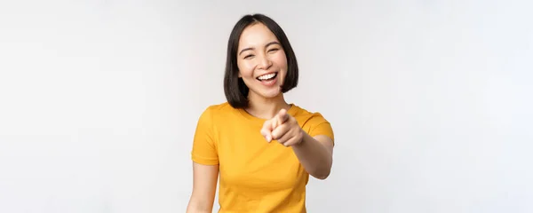 快乐美丽的亚洲女人笑着，手指指向相机，咯咯地笑着，无忧无虑地笑着，穿着黄色T恤站在白色的背景上 — 图库照片