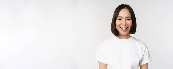 De cerca retrato de joven mujer asiática mirando a la cámara, vistiendo camiseta, sonriendo y mirando feliz, fondo blanco —  Fotos de Stock