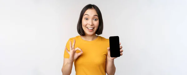 Aufgeregte Asiatin zeigt Handybildschirm, Okay-Schild, empfiehlt Smartphone-App, steht in gelbem T-Shirt vor weißem Hintergrund — Stockfoto