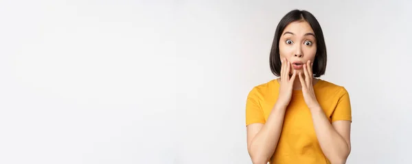 Close up portrait of asian woman looking surprised, wow face, staring impressed at camera, standing over white background in yellow t-shirt — Stock Photo, Image