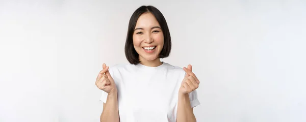 Hermosa mujer asiática sonriendo, mostrando el gesto de los corazones de los dedos, usando camiseta, de pie sobre fondo blanco — Foto de Stock