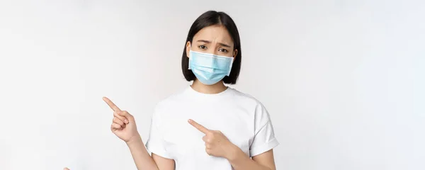 Sorgsen asiatisk kvinna i medicinsk mask, pekar finger vänster, rynkar pannan och ser upprörd, klagar, visar banner, står över vit bakgrund — Stockfoto