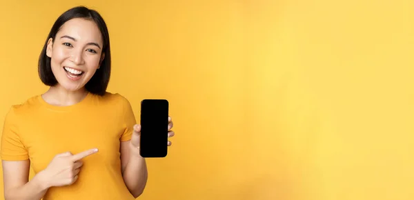 Mooi gelukkig aziatisch meisje tonen mobiele telefoon scherm, toepassing op smartphone gadget, staande over gele achtergrond — Stockfoto