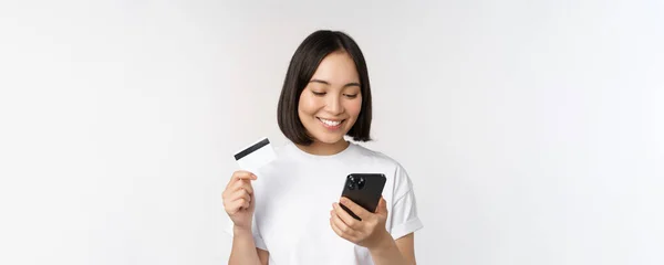 Compras online. Feliz mulher asiática usando cartão de crédito e aplicativo de smartphone, pagando no site via telefone celular, fundo branco — Fotografia de Stock