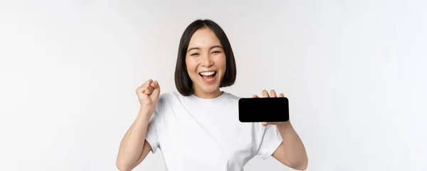 Entusiástico asiático menina gritar de alegria, mostrando tela de smartphone horizontal, exibição de telefone celular, de pé sobre fundo branco — Fotografia de Stock