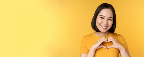 Κοντινό πλάνο πορτρέτο της χαμογελαστής Κορεάτισσας γυναίκας, που δείχνει ρομαντικό σημάδι της καρδιάς και φαίνεται ευτυχισμένη, στέκεται πάνω από κίτρινο φόντο — Φωτογραφία Αρχείου