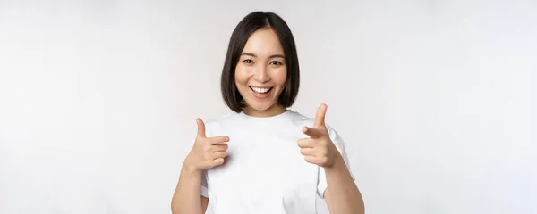 Imagen de chica asiática sonriente apuntando con los dedos a la cámara, eligiendo, invitándote, felicitando, de pie en camiseta sobre fondo blanco —  Fotos de Stock