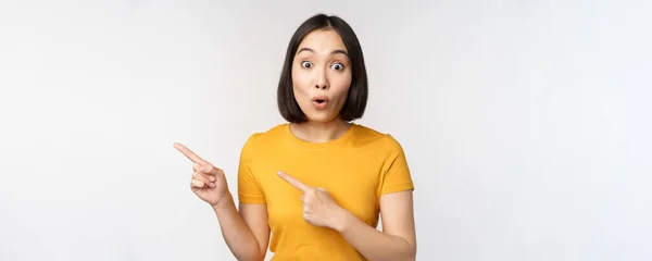 Portret van lachend aziatisch brunette meisje in geel T-shirt, wijzende vingers links, tonen kopieerruimte, promo deal, demonstreren banner, staande over witte achtergrond — Stockfoto