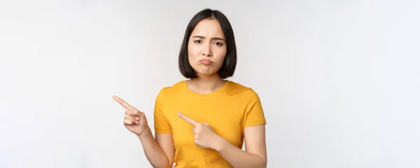 Retrato de morena decepcionada chica asiática, señalando los dedos a la izquierda, mutilando molesto, mostrando smth desagradable, de pie sobre fondo blanco —  Fotos de Stock