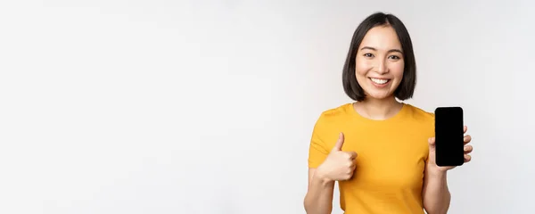 Technologie und Menschen. Lächelnde junge Frau zeigt Daumen hoch und Smartphone-Bildschirm, Handy-App-Schnittstelle, steht vor weißem Hintergrund — Stockfoto