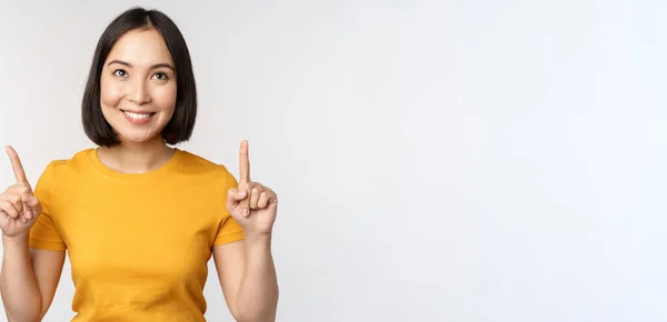 Glückliche schöne asiatische Mädchen zeigt Werbung, zeigt mit den Fingern nach oben, steht über weißem Hintergrund — Stockfoto