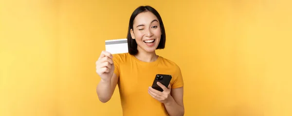 Chica asiática alegre sonriendo, mostrando la tarjeta de crédito y el teléfono inteligente, recomendando la banca del teléfono móvil, de pie sobre el fondo amarillo —  Fotos de Stock