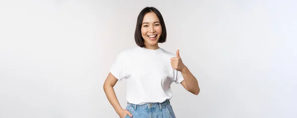 Glad vacker koreansk kvinna, leende nöjd, visar tummen upp i godkännande, rekommendera varumärke eller företag, står över vit bakgrund — Stockfoto