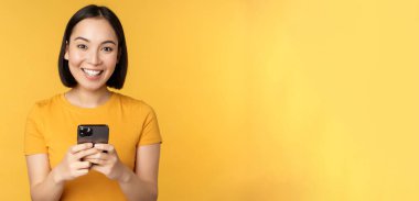 Teknoloji. Gülümseyen Asyalı kadın cep telefonu kullanıyor, akıllı telefonu elinde tutuyor, sarı arka planda tişörtle dikiliyor.