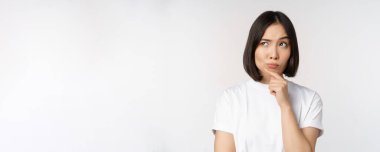 Düşünceli Asyalı kadın yana bakıyor, düşünüyor, varsayıyor, düşünüyor ya da smth 'yi seçiyor, beyaz arka planda duruyor.