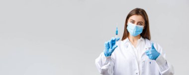 Covid-19, virüsü önleme, sağlık çalışanları ve karantina konsepti. Tıbbi maske ve lastik eldiven takmış kararlı genç doktor, önlükler baş parmak ve şırıngada koronavirüs aşısı gösteriyor.