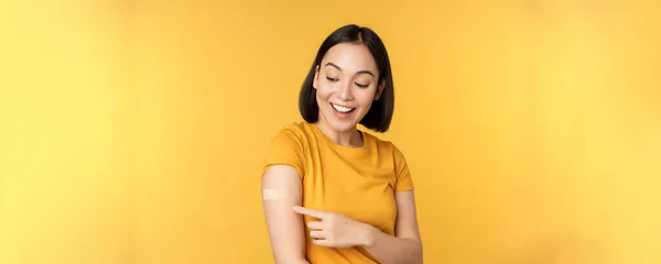 Vacinação e conceito de pandemia de covid-19. Feliz e saudável menina asiática apontando para o ombro com ajuda banda depois de vacinar de coronavírus, fundo amarelo — Fotografia de Stock