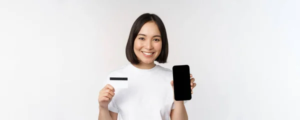 Portret van lachende jonge aziatische vrouw tonen mobiele telefoon scherm en creditcard, staande over witte achtergrond — Stockfoto