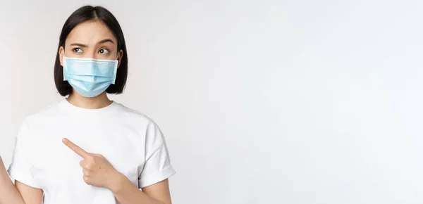 Imagem de mulher asiática em máscara médica de covid, olhando confuso à esquerda, apontando para o logotipo, mostrando propaganda, de pé sobre fundo branco — Fotografia de Stock