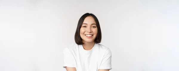 Beautiful korean girl smiling, white teeth, looking lovely at camera, standing in white tshirt over studio background — Stock Photo, Image