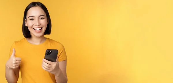 Glad leende asiatisk flicka som håller mobiltelefon och visar tummen upp, rekommendera ansökan på smartphone, står över gul bakgrund — Stockfoto