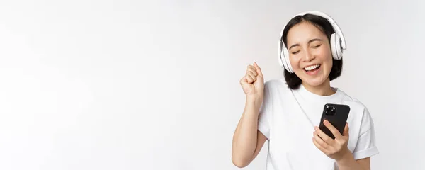 Belle fille asiatique moderne, écouter de la musique dans les écouteurs, tenant le téléphone mobile, en utilisant l'application smartphone, debout sur fond blanc — Photo