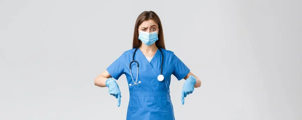 Covid-19, preventing virus, health, healthcare workers and quarantine concept. Skeptical and unsure female nurse or doctor in blue scrubs, personal protective equipment, pointing fingers down unsure — Stock Photo, Image