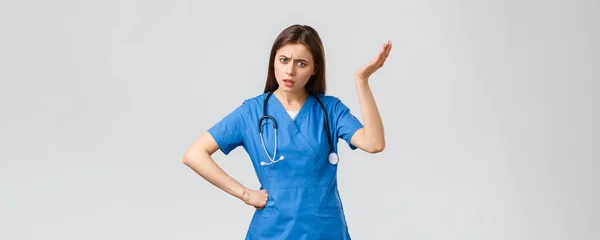 Pracownicy medyczni, opieka zdrowotna, koncepcja covid-19 i szczepień. Sfrustrowana i zdezorientowana młoda pielęgniarka, piękna lekarka w niebieskim fartuchu, słyszy dziwny pomysł, wzrusza ręką podniesioną nonsensem — Zdjęcie stockowe