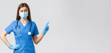 Covid-19, virüsün önlenmesi, sağlık, sağlık çalışanları ve karantina konsepti. Şüpheci ve hoşnutsuz kadın hemşire veya doktor mavi önlüklü, tıbbi maskeli, sağı gösteren, kaşlarını çatan