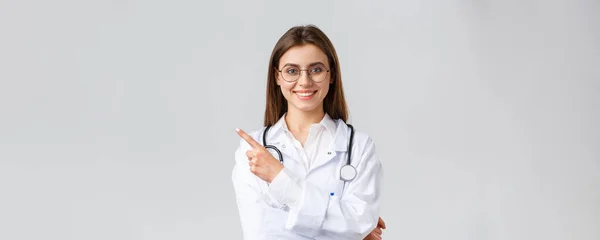 Pracownicy służby zdrowia, medycyna i koncepcja pandemii covid-19. Dobrze wyglądająca profesjonalna lekarka w białym fartuchu i okularach, wytykająca palcami w lewo, uśmiechnięta przyjazna, pokazująca informacje — Zdjęcie stockowe