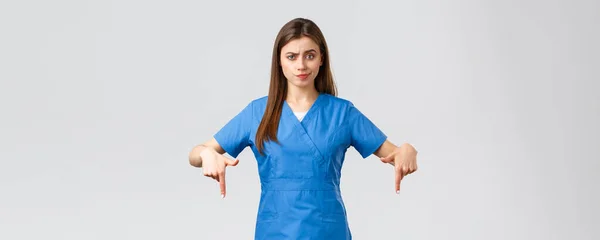 Gesundheitshelfer, Virusprävention, Covid-19-Test-Screening, Medikamentenkonzept. Skeptische, enttäuschte Krankenschwester oder Ärztin im blauen Peeling zeigt mit dem Finger nach unten, lächelt unzufrieden, ist zweifelhaft — Stockfoto