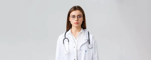 Sjukvårdspersonal, medicin, försäkring och covid-19 pandemi koncept. Allvarligt utseende smart professionell läkare, sjuksköterska i vita skurar och stetoskop, utseende kamera bestäms, grå bakgrund — Stockfoto